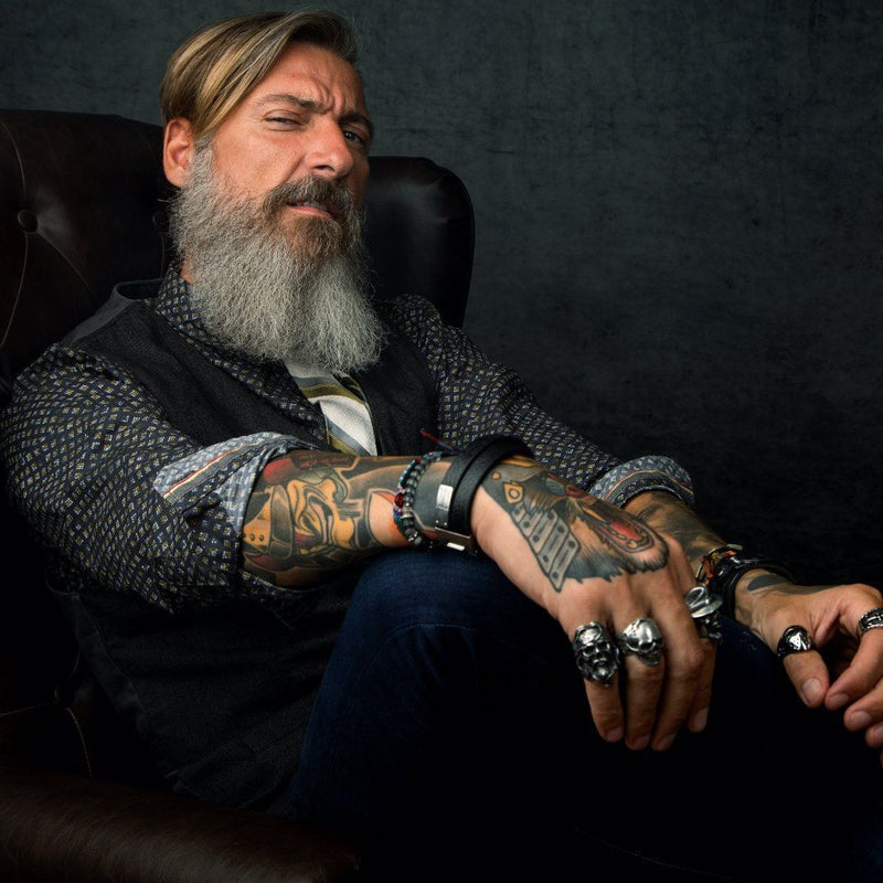 Close-up of older man with brown hair and grey beard seated in leather chair looking at the camera. Gentleman is well dressed and has sleeve tattoos and is wearing much jewelry